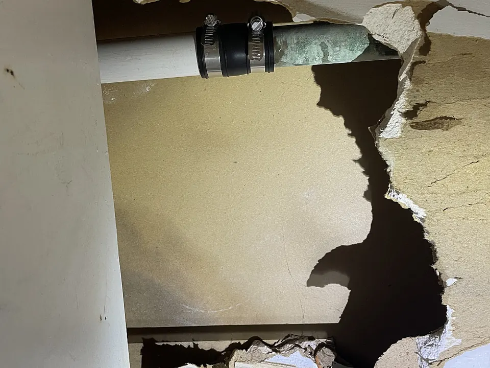 Torn, water-damaged sheetrock behind an old metal drain pipe under a bathroom sink